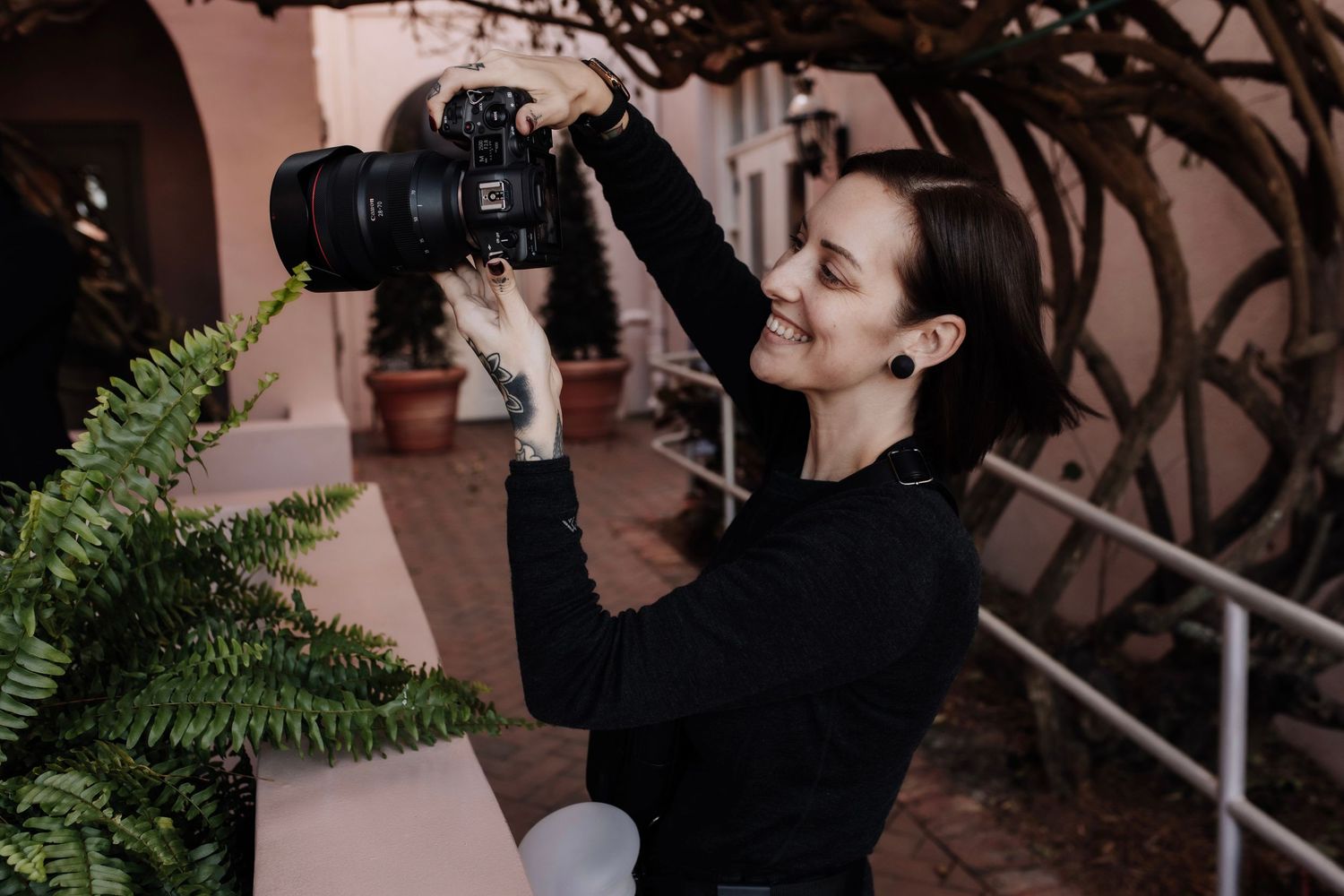 Smiling woman in black turtleneck holds DSLR camera outdoors near fern plants and pink building.