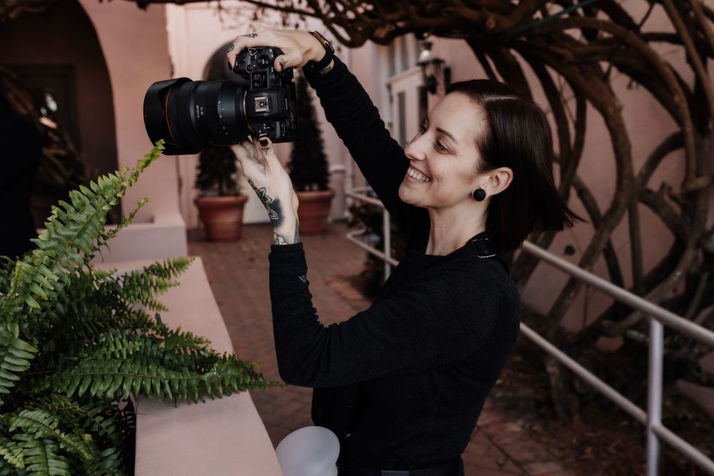 Smiling woman in black turtleneck holds DSLR camera outdoors near fern plants and pink building.