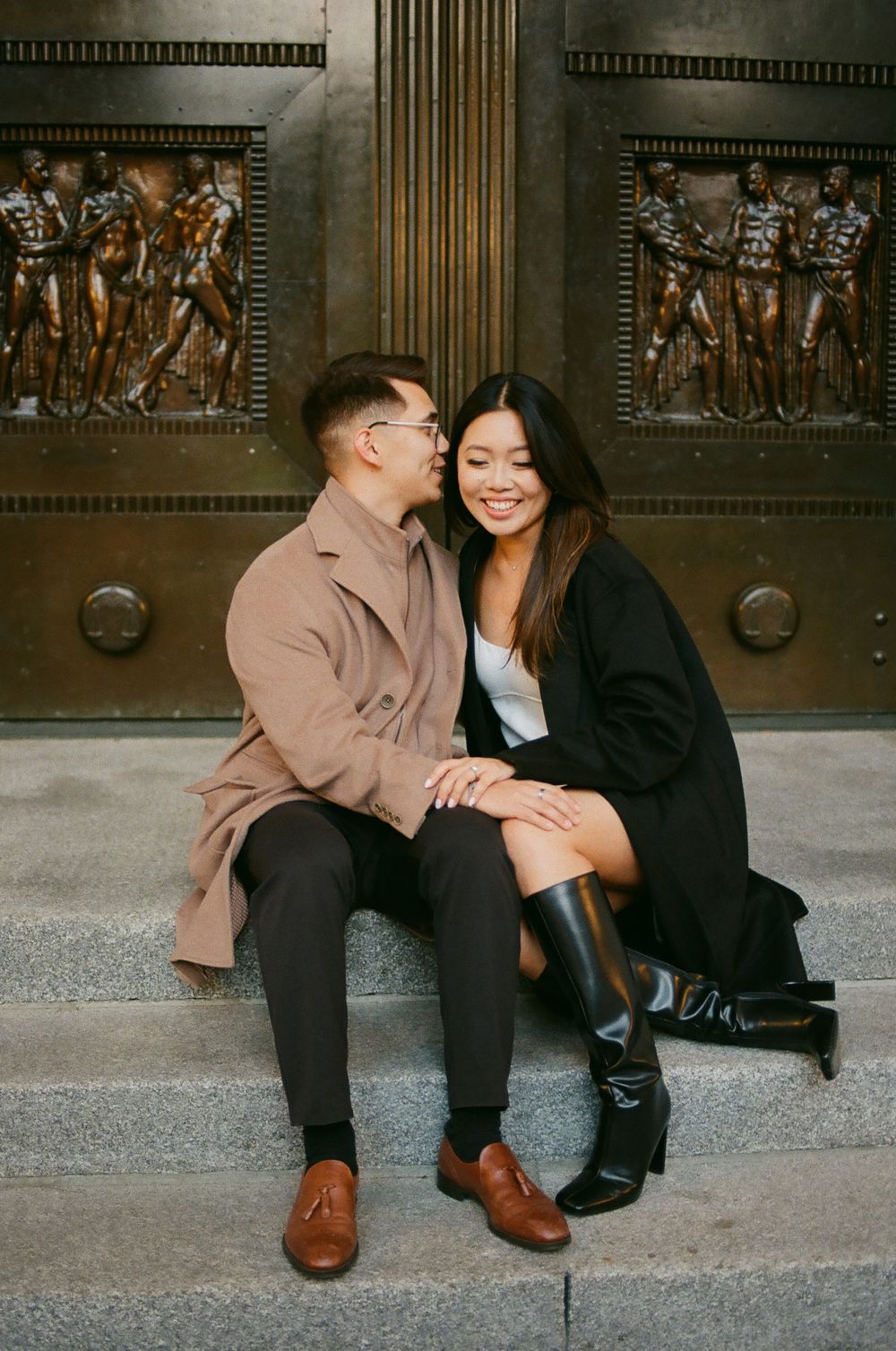 Couple sitting on steps before ornate bronze doors, sharing a tender moment in coordinating coats. Film wedding photographer Aliza Dworkind