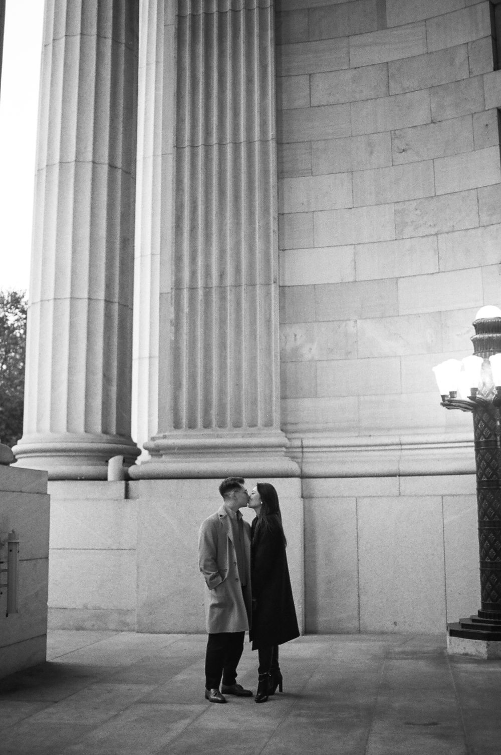Couple sharing a romantic kiss between grand stone columns in a black and white photo. Aliza Dworkind photographer in Montreal