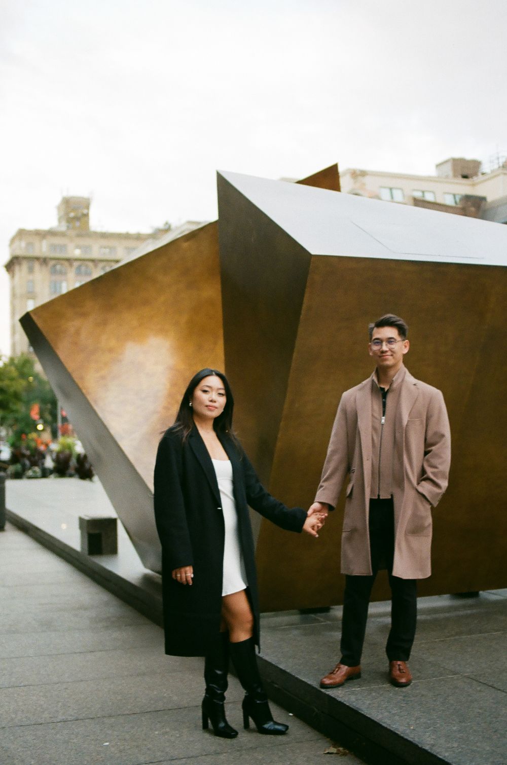 Couple holding hands in front of a large geometric bronze sculpture in an urban plaza, dressed in elegant long coats. Montreal Old Port film