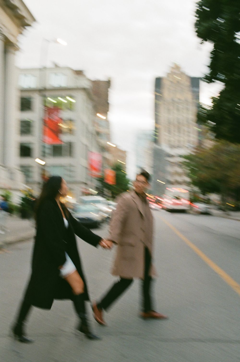 Couple holding hands crossing a city street, wearing long coats in black and tan, with urban buildings in the background. 35mm film photos