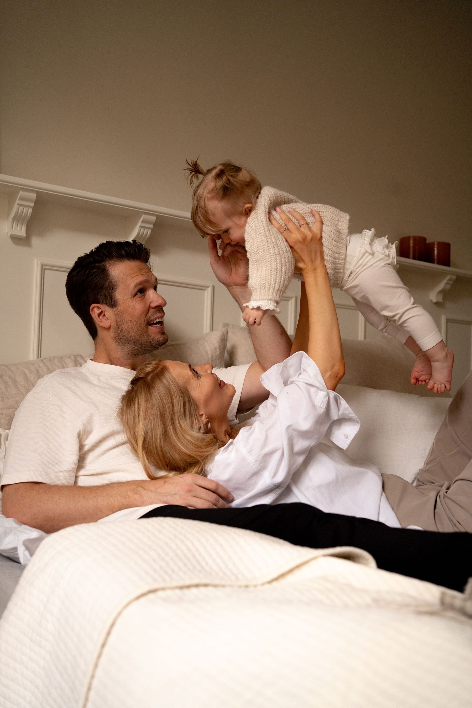 En mamma och pappa ligger i en säng. Mamman ligger mot pappans bröst och håller upp sitt barn som flyger ovanför dem.
