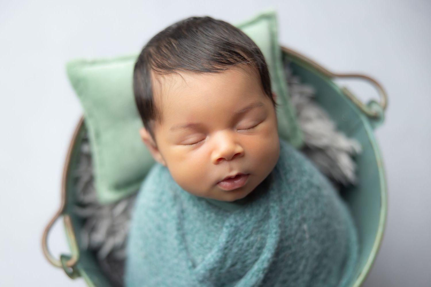 Sleeping newborn baby wrapped in teal swaddle inside a small bucket with a green pillow.