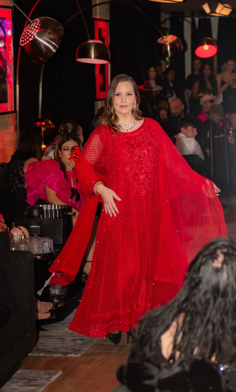 Model in flowing red sparkly gown walking the runway at a fashion show.