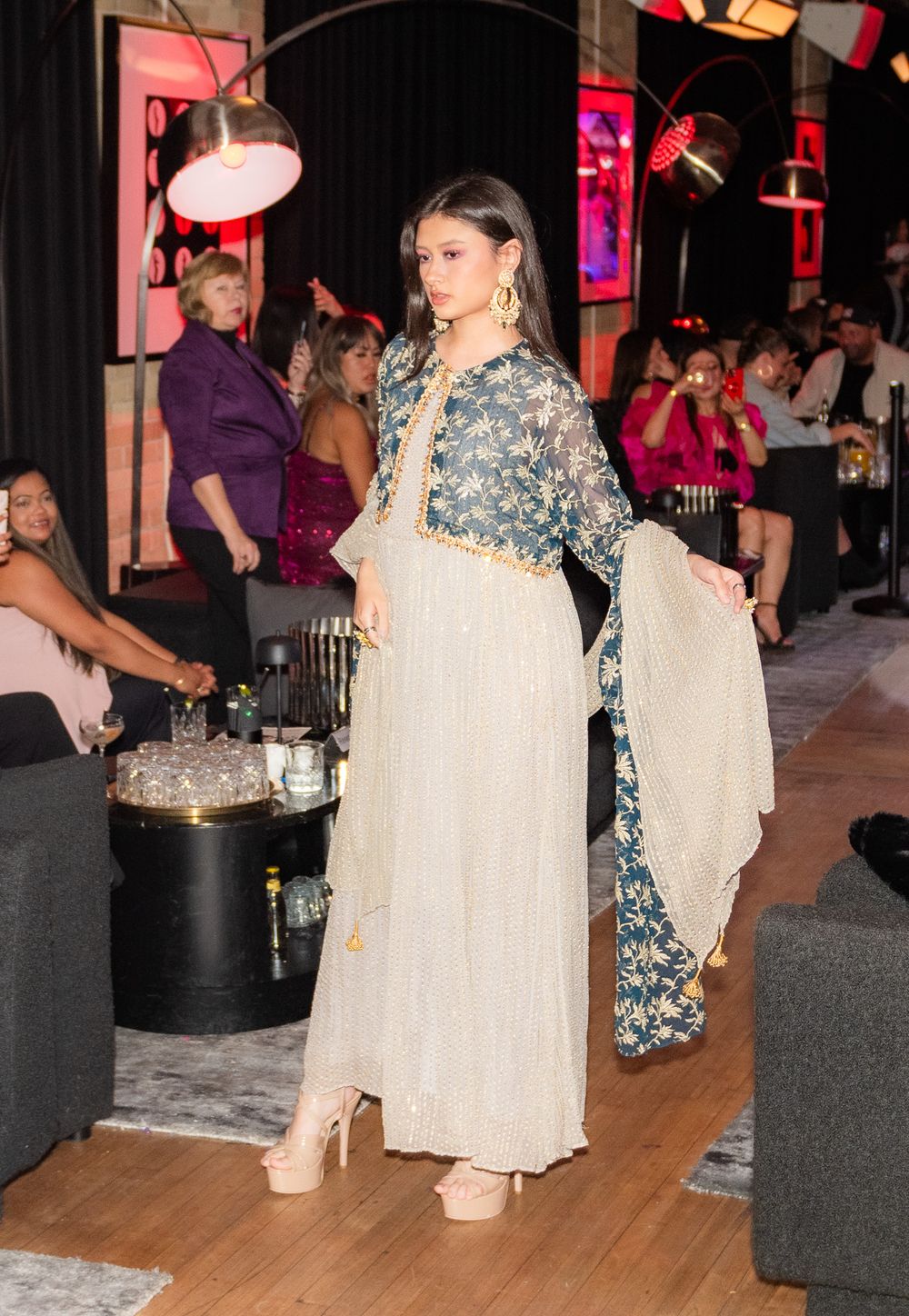 Fashion model wearing cream and blue patterned dress on runway with dramatic lighting.