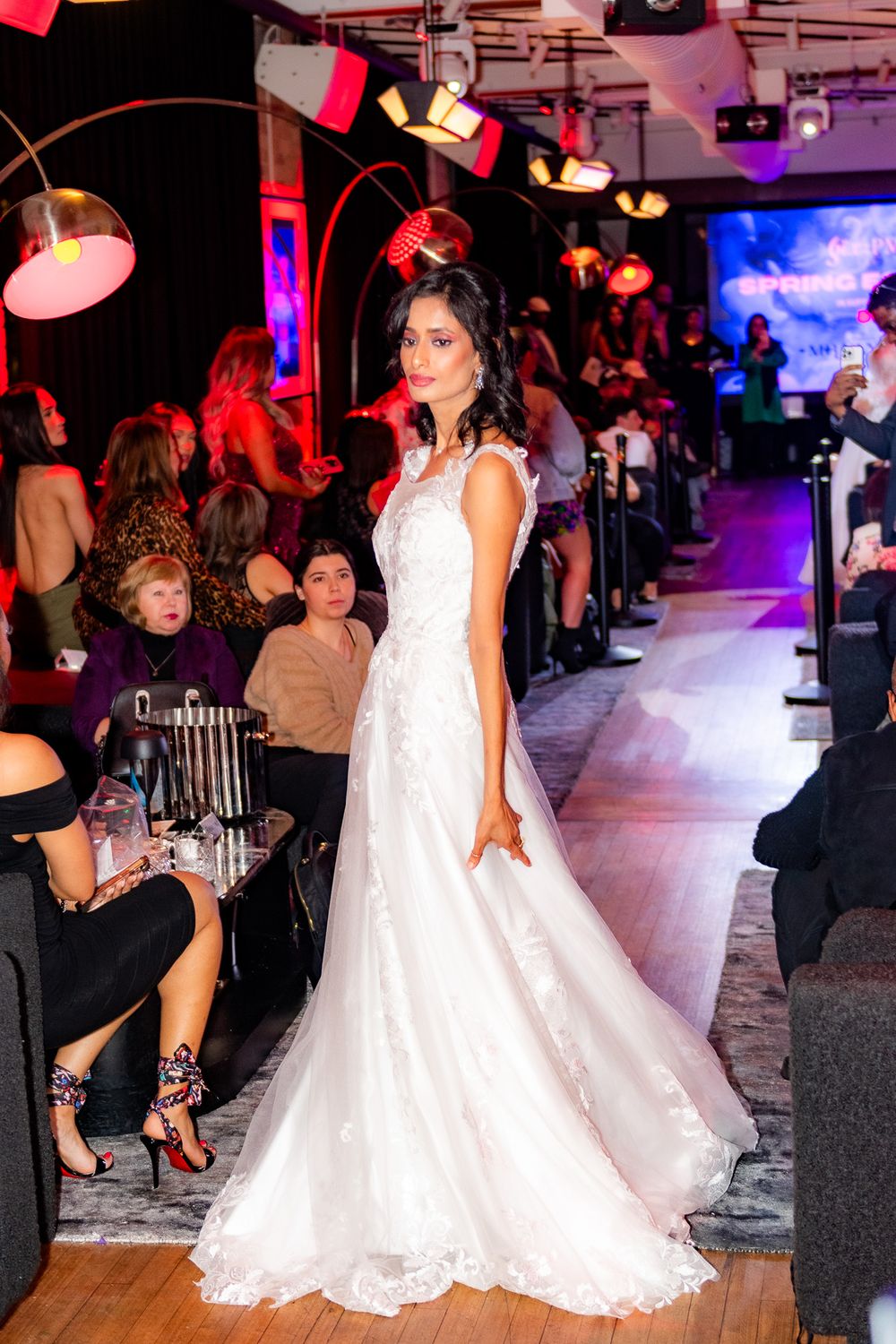 Model in white lace wedding gown walking runway under dramatic red lighting.