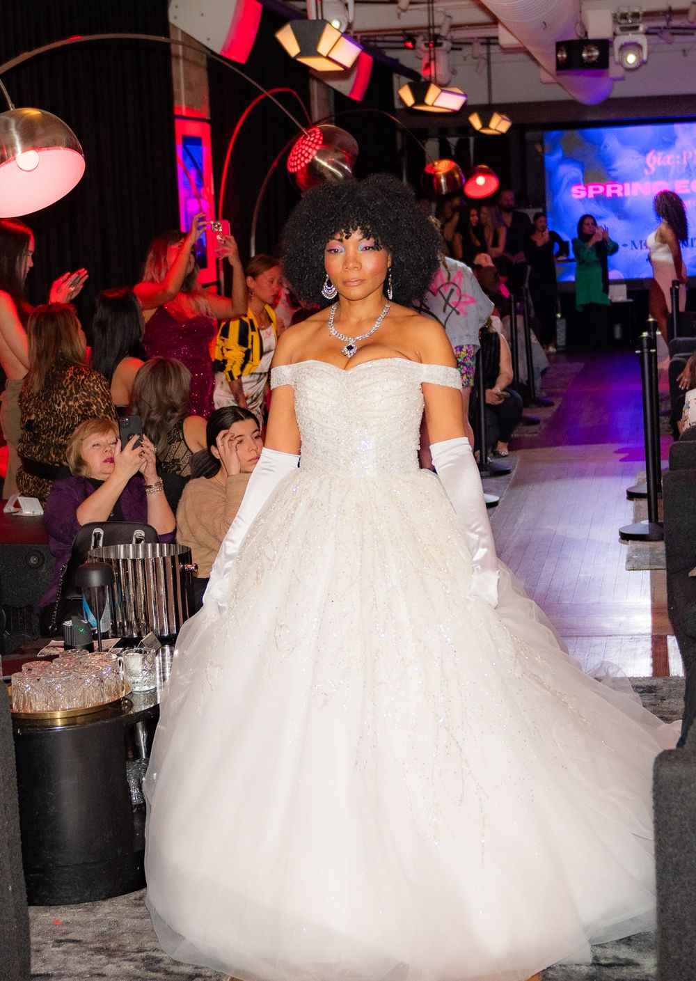 Model in princess-style white wedding gown and gloves on fashion runway.