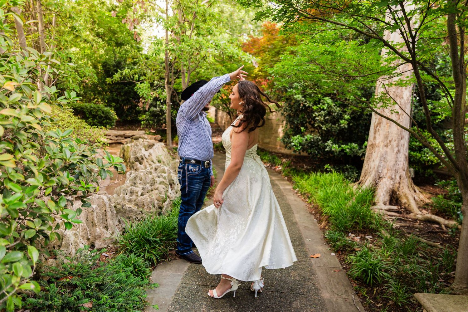 Bride in white dress twirls with groom on garden path surrounded by lush green trees and foliage.