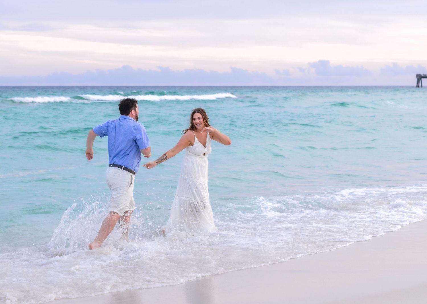 Couple holding hands running through turquoise ocean waves on a tropical white sand beach at sunset.