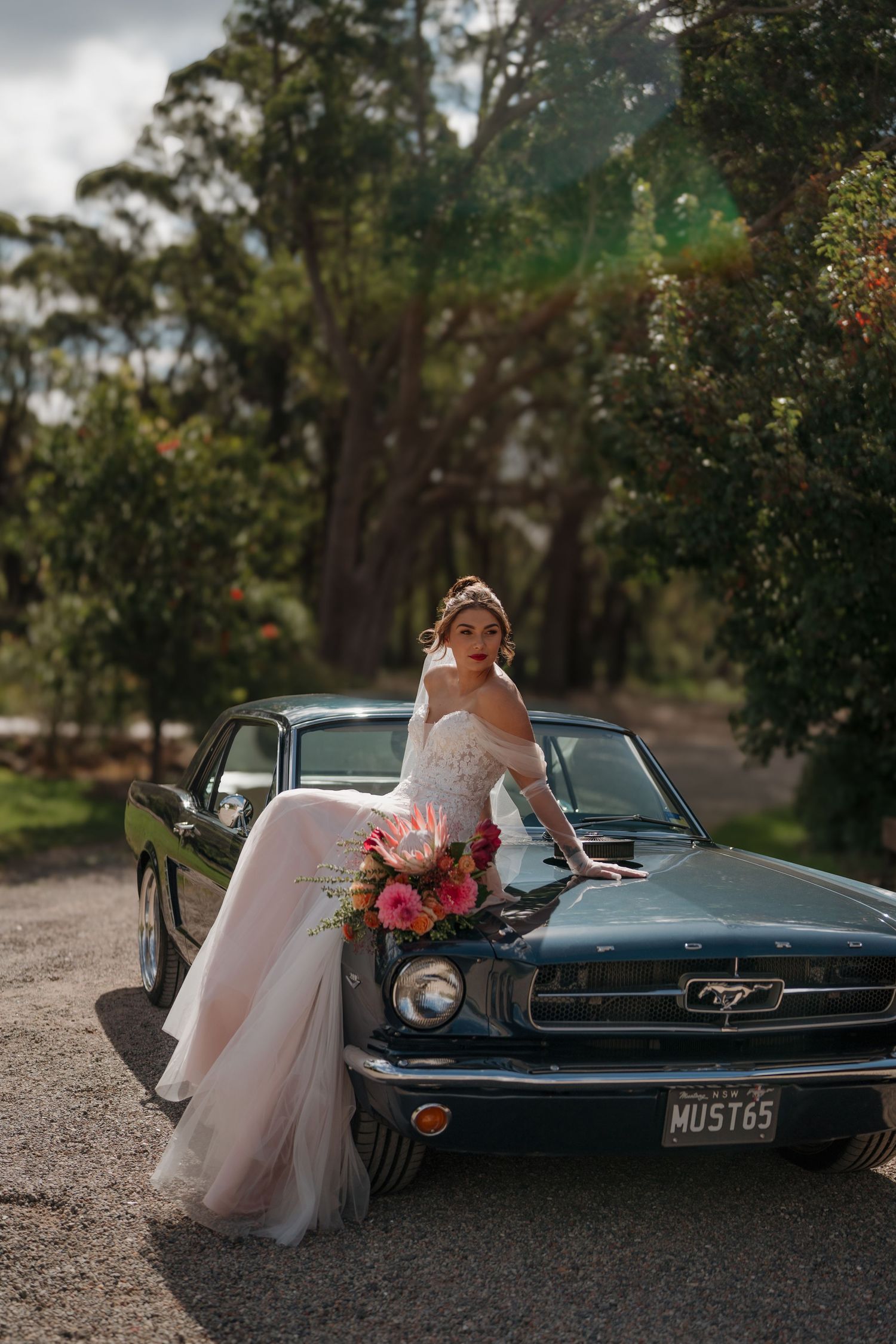 Bride in white gown leaning on a classic Ford Mustang with pink florals, surrounded by lush green trees.