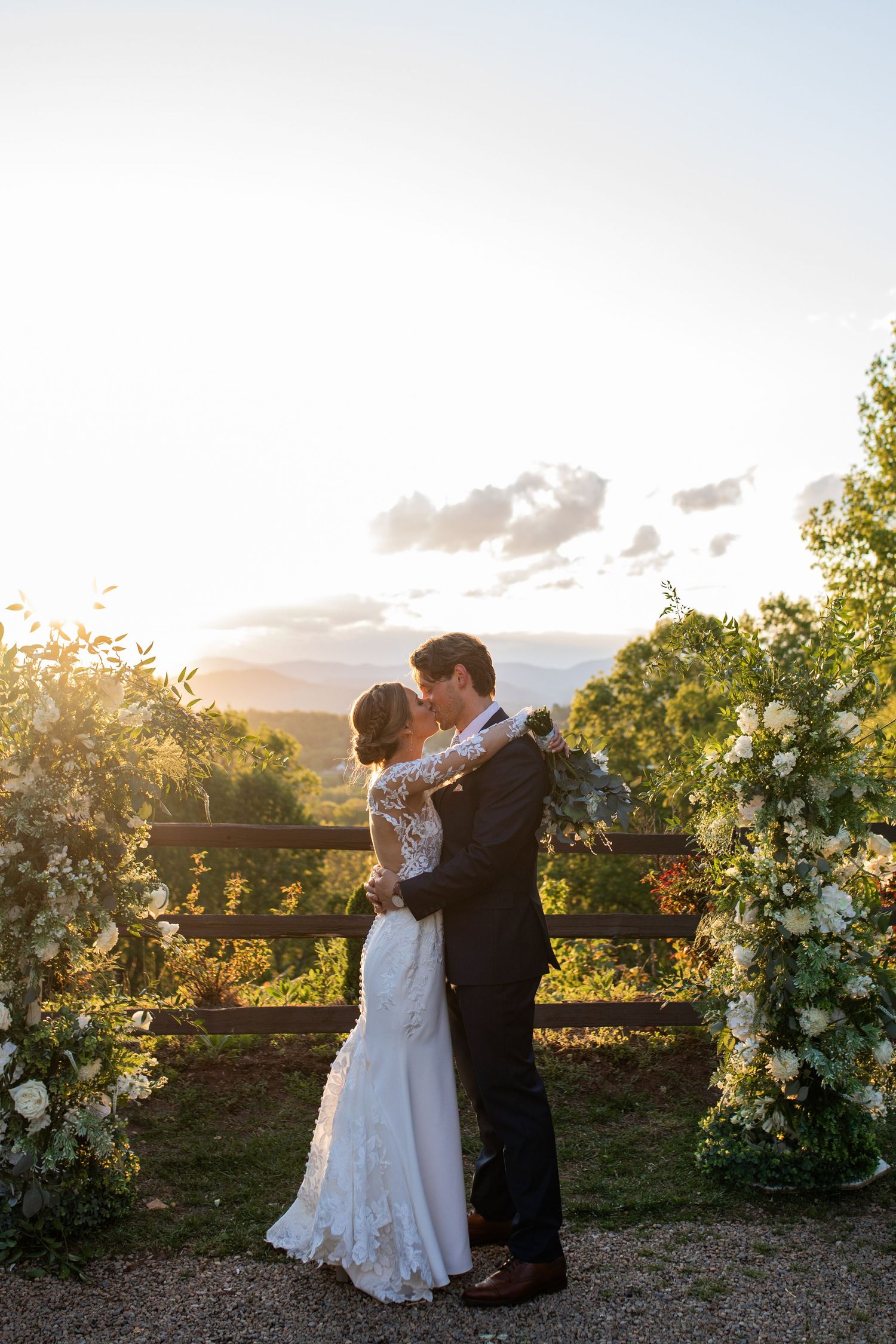 Bride and groom embrace at sunset during outdoor wedding ceremony with white floral arrangements and mountain views.