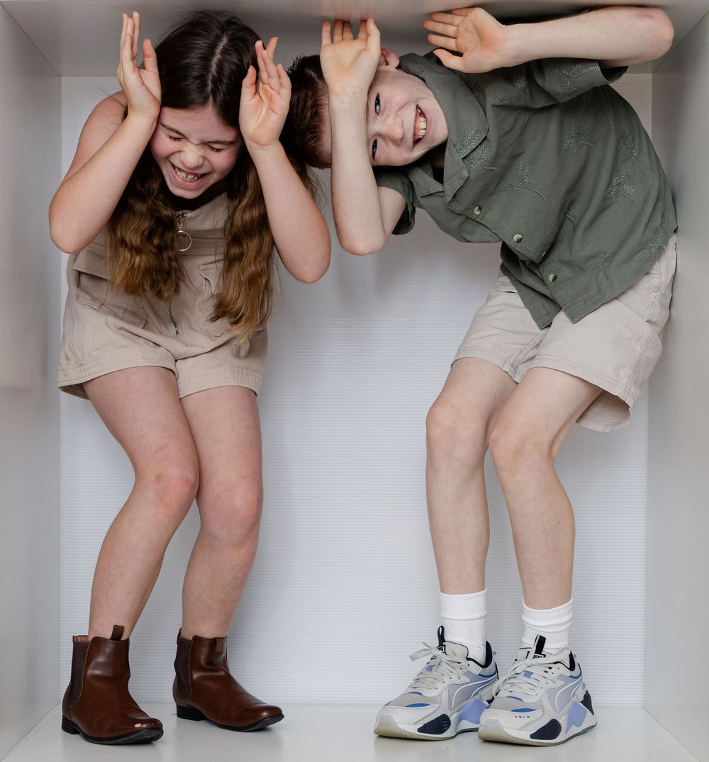 Two playful kids laughing and making silly faces with hands on heads, wearing casual outfits and sneakers.