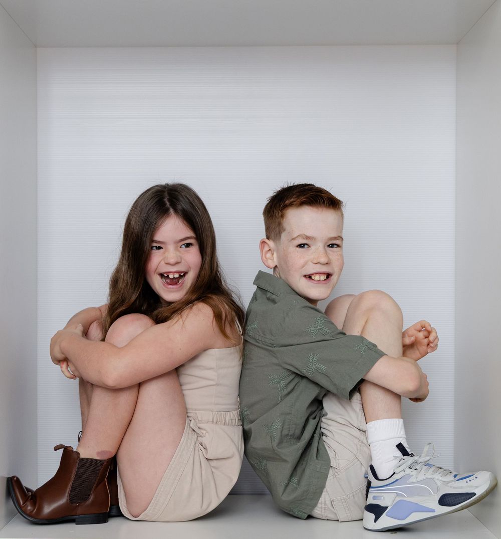Two happy children sitting back-to-back on a white shelf, smiling at the camera.