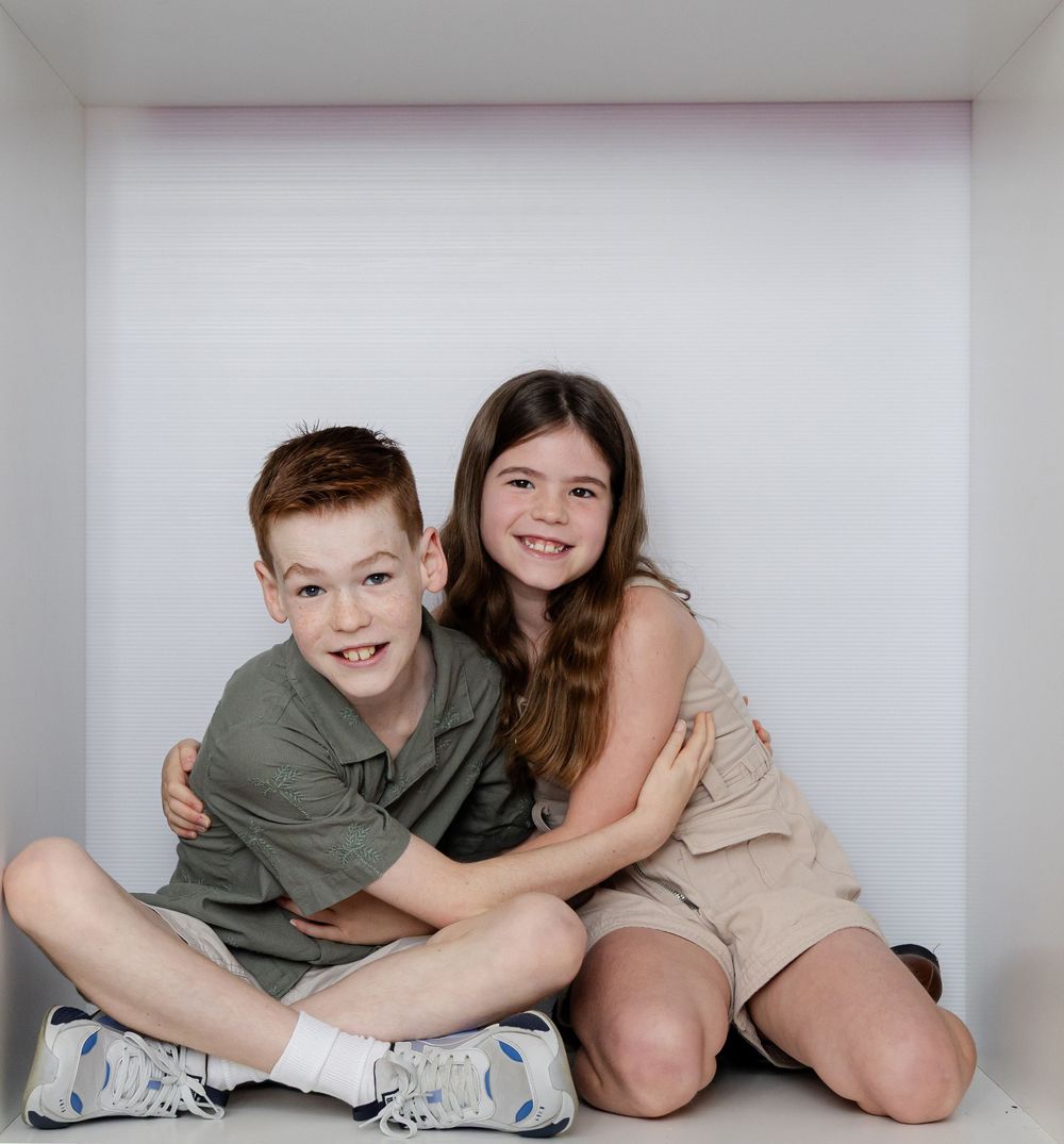 Two smiling children, a red-haired boy and a brunette girl, pose together in a white box setting.