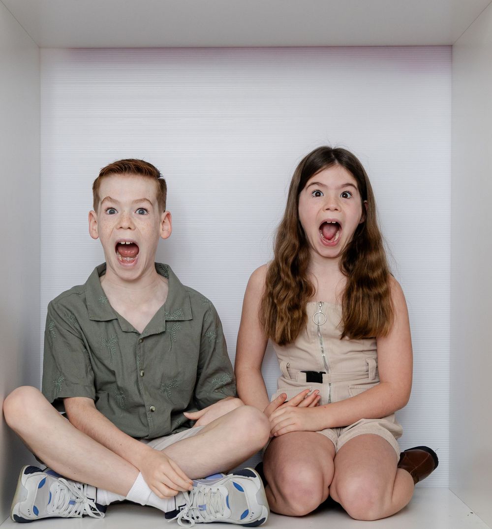 Two kids sitting cross-legged in a white box, mouths open wide in surprised expressions, wearing casual outfits.