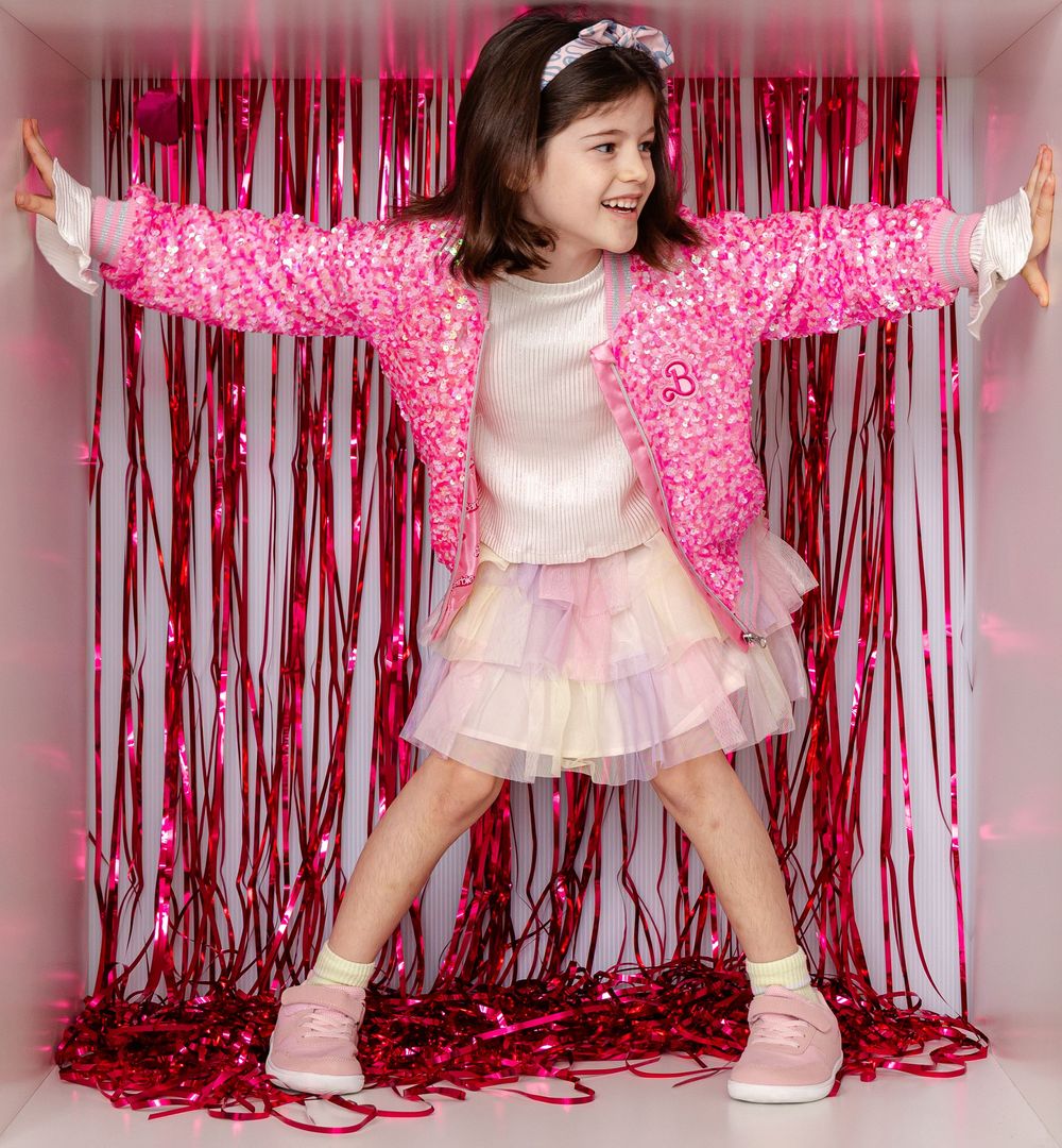 Young girl in pink sequin jacket and tutu skirt poses with arms wide in a shimmery pink tinsel curtain backdrop.