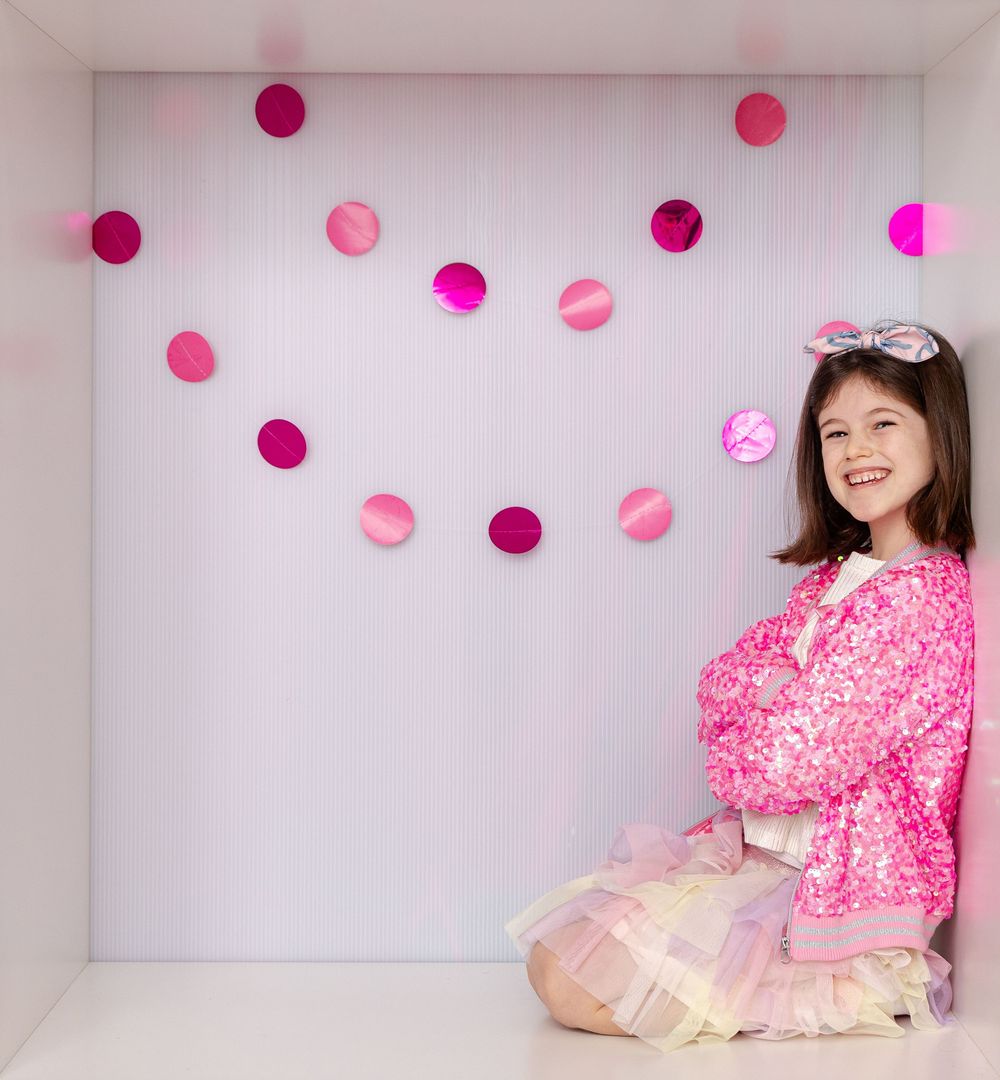 Smiling girl in pink sequin jacket and rainbow tutu sits cross-legged beneath pink circle garland decorations.