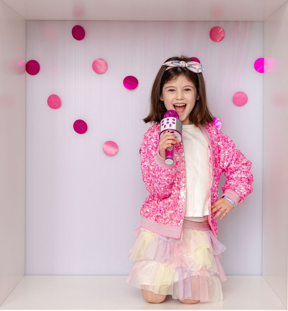 A cheerful girl in a pink sequin jacket sings into a microphone surrounded by pink confetti circles.