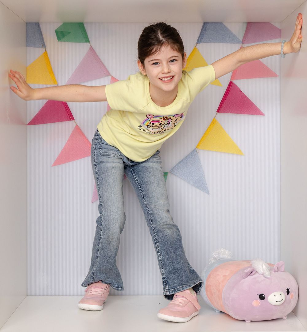 Girl in yellow tee and jeans poses with arms outstretched in a colorful bunting-decorated white box with a plush toy.
