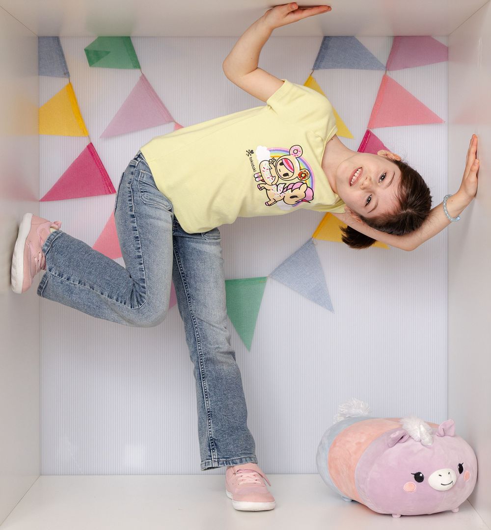 A playful girl in yellow tee and jeans poses sideways in a colorful geometric box with a pink stuffed animal.