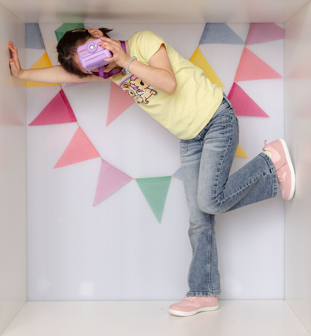 Young girl holding purple camera poses dynamically in a festive box with colorful triangle bunting.