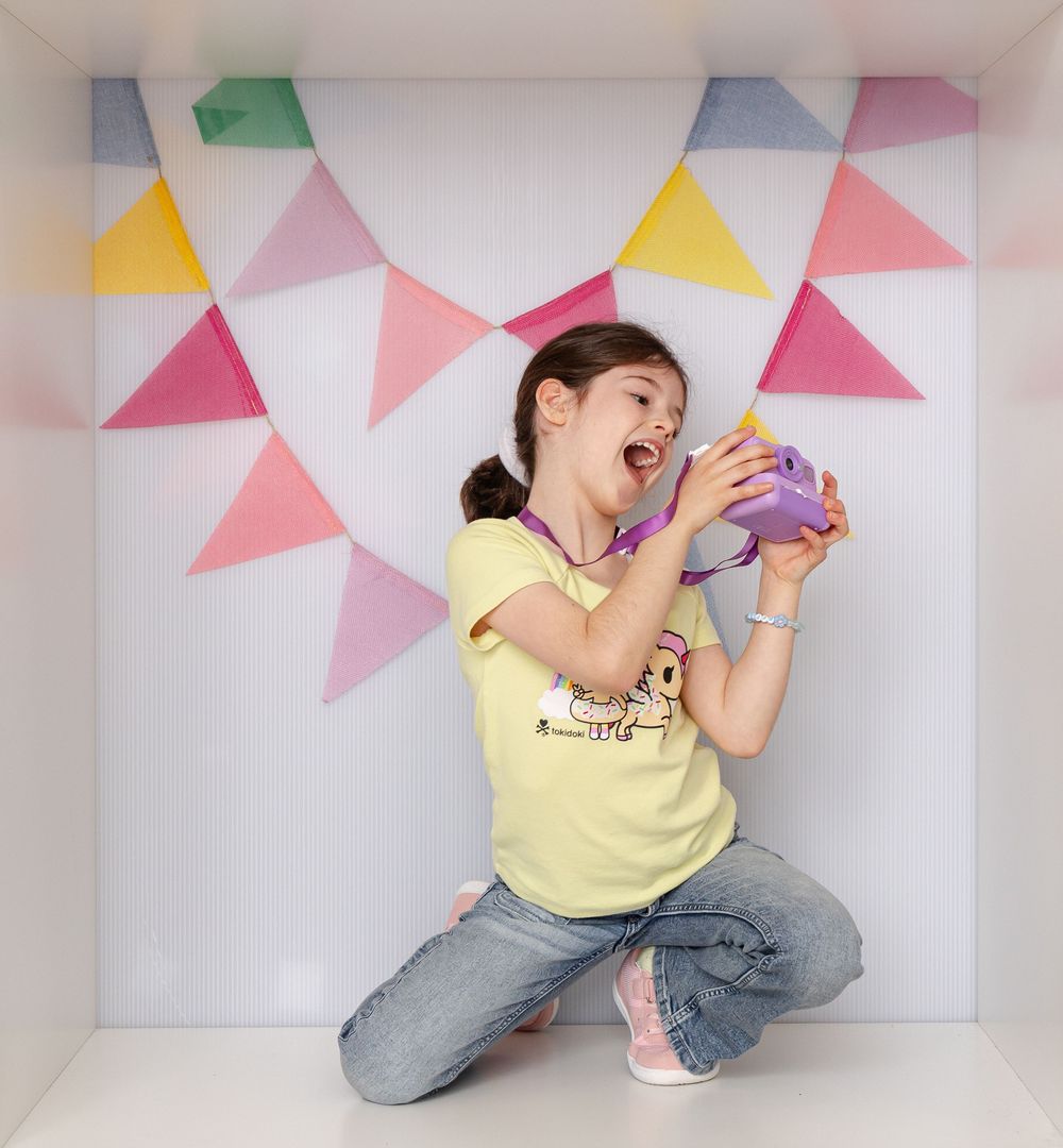 An excited girl in a yellow graphic tee holds a purple toy camera inside a colorful bunting-decorated white box.