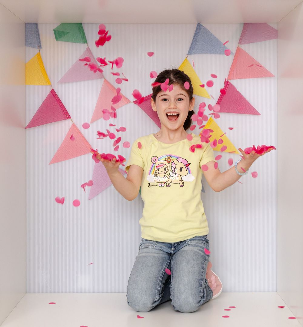 Excited girl in yellow graphic tee kneels as pink confetti falls around her in a festive bunting-decorated space.