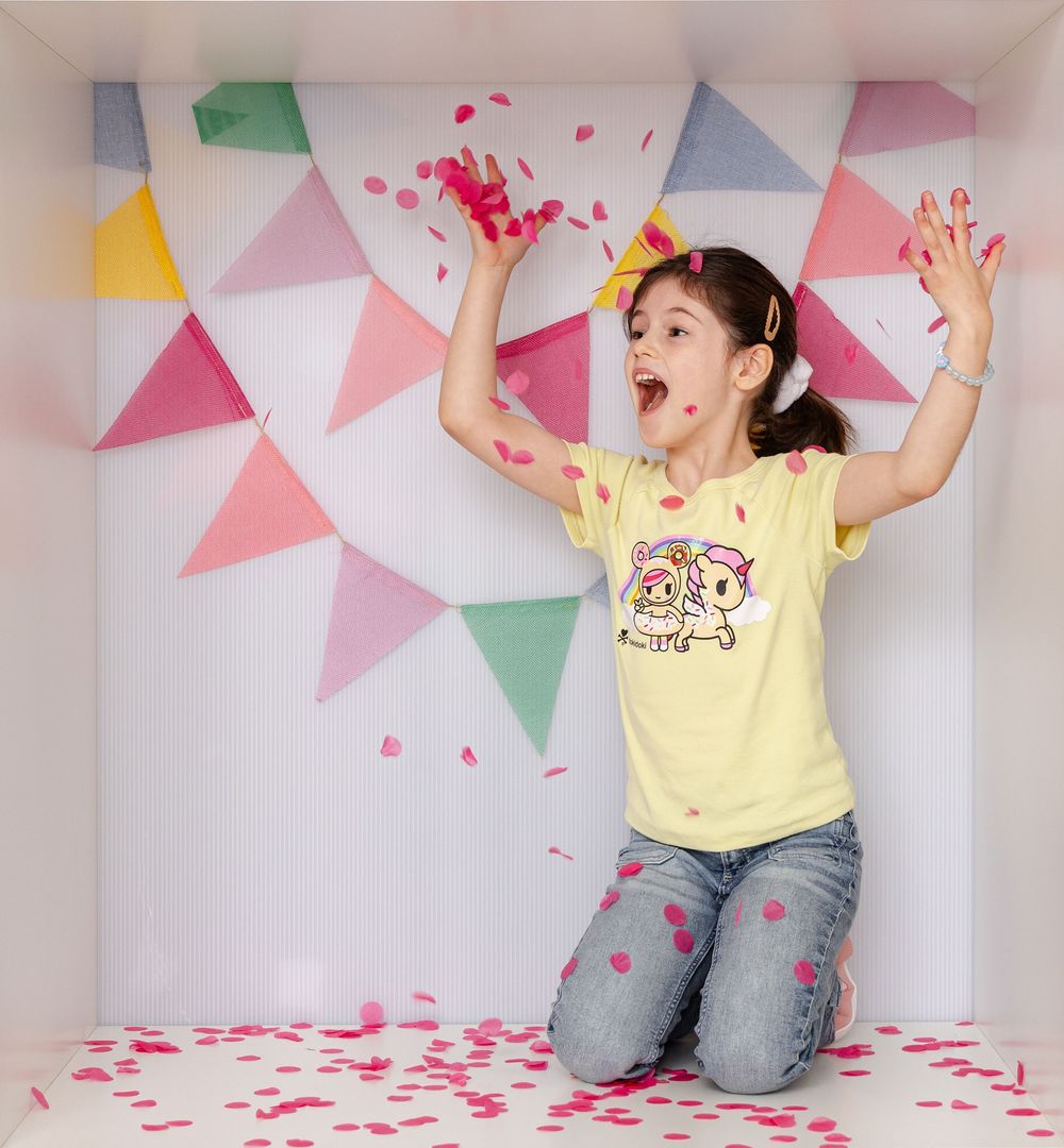 Excited girl kneeling as pink confetti falls around her in a bright, bunting-decorated photo box.