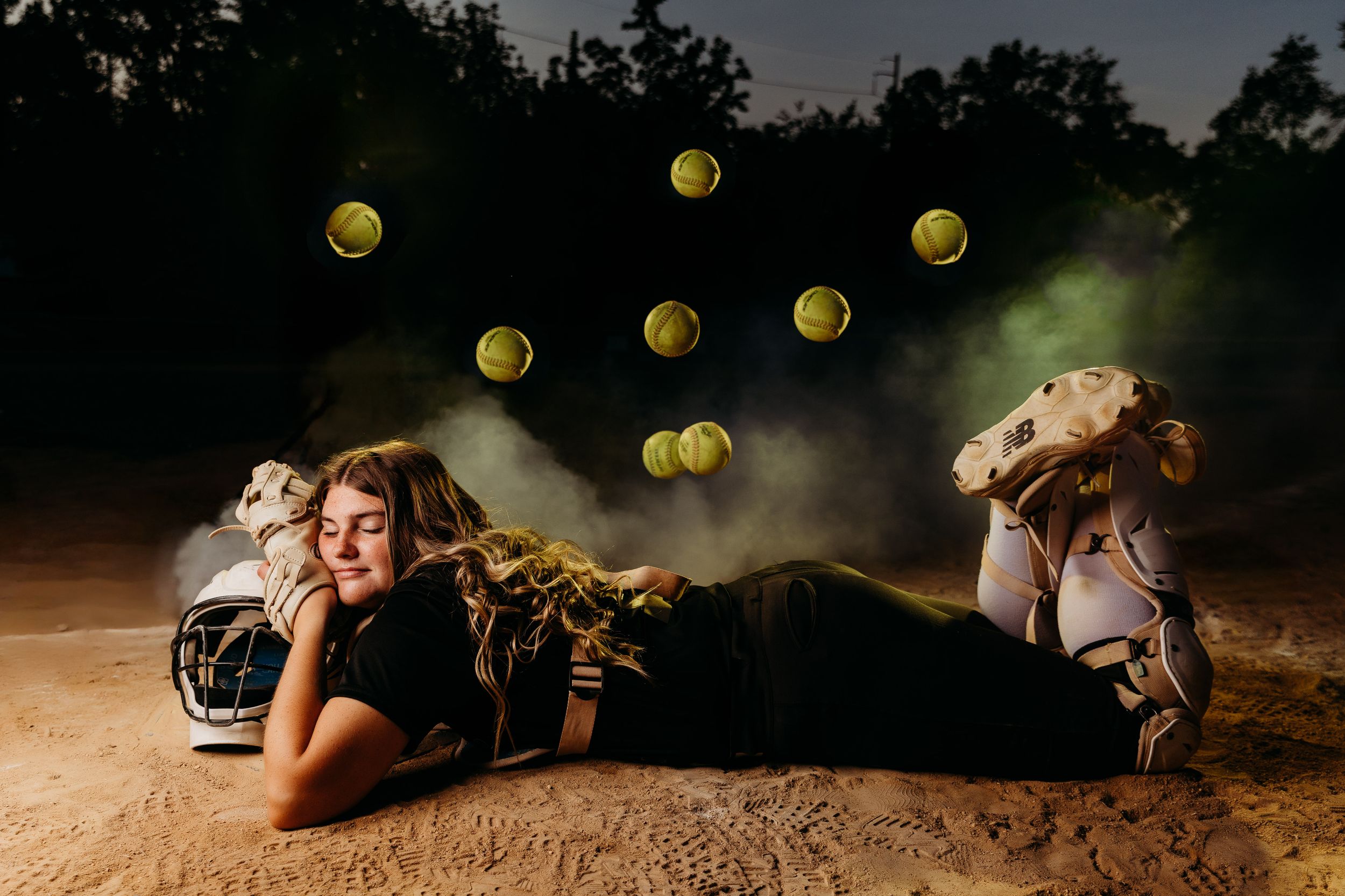 Softball player lying on dirt field with tennis balls floating in smoky air, wearing cleats and black uniform at night.
