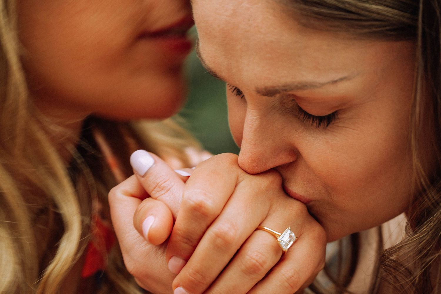 Two women share a tender moment as one kisses the other's hand adorned with a diamond engagement ring. After secret proposal moment