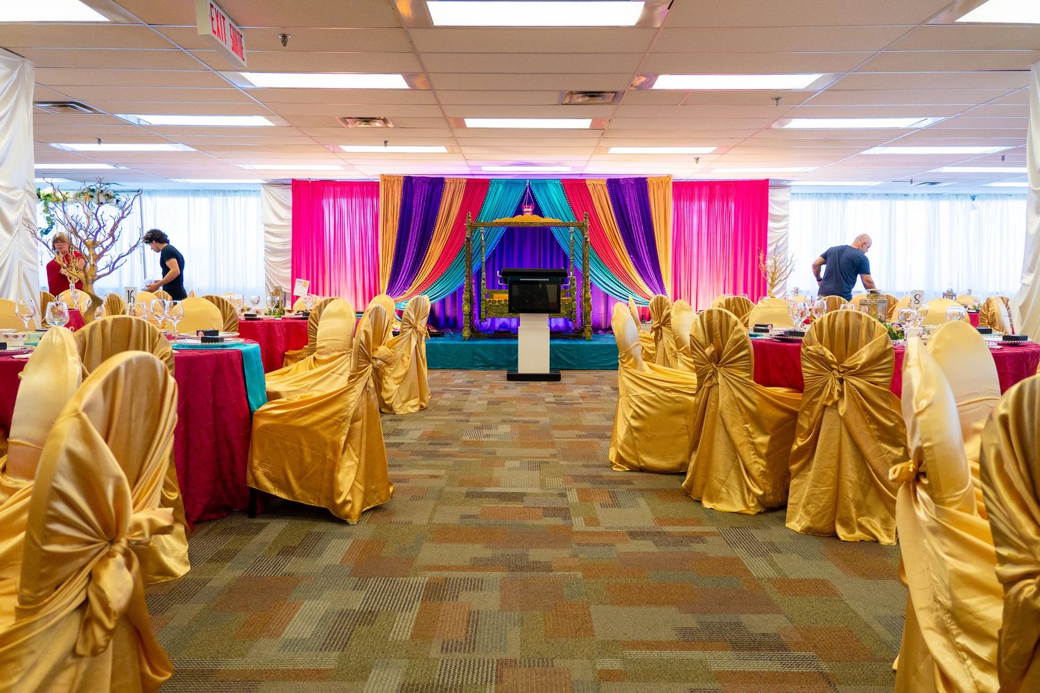 Elegant banquet hall decorated with colorful drapes, gold chair covers, and vibrant pink and teal stage backdrop for a festive event.