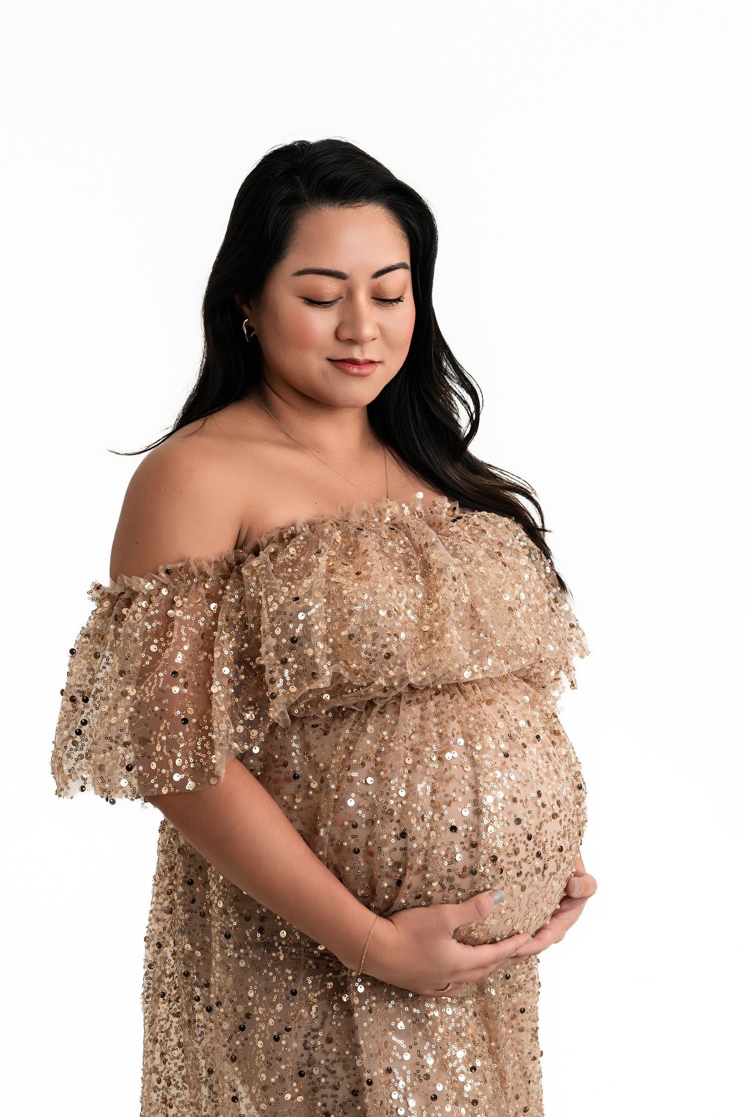 Pregnant woman in gold sequin off-shoulder gown cradling baby bump, studio maternity portrait on white background.