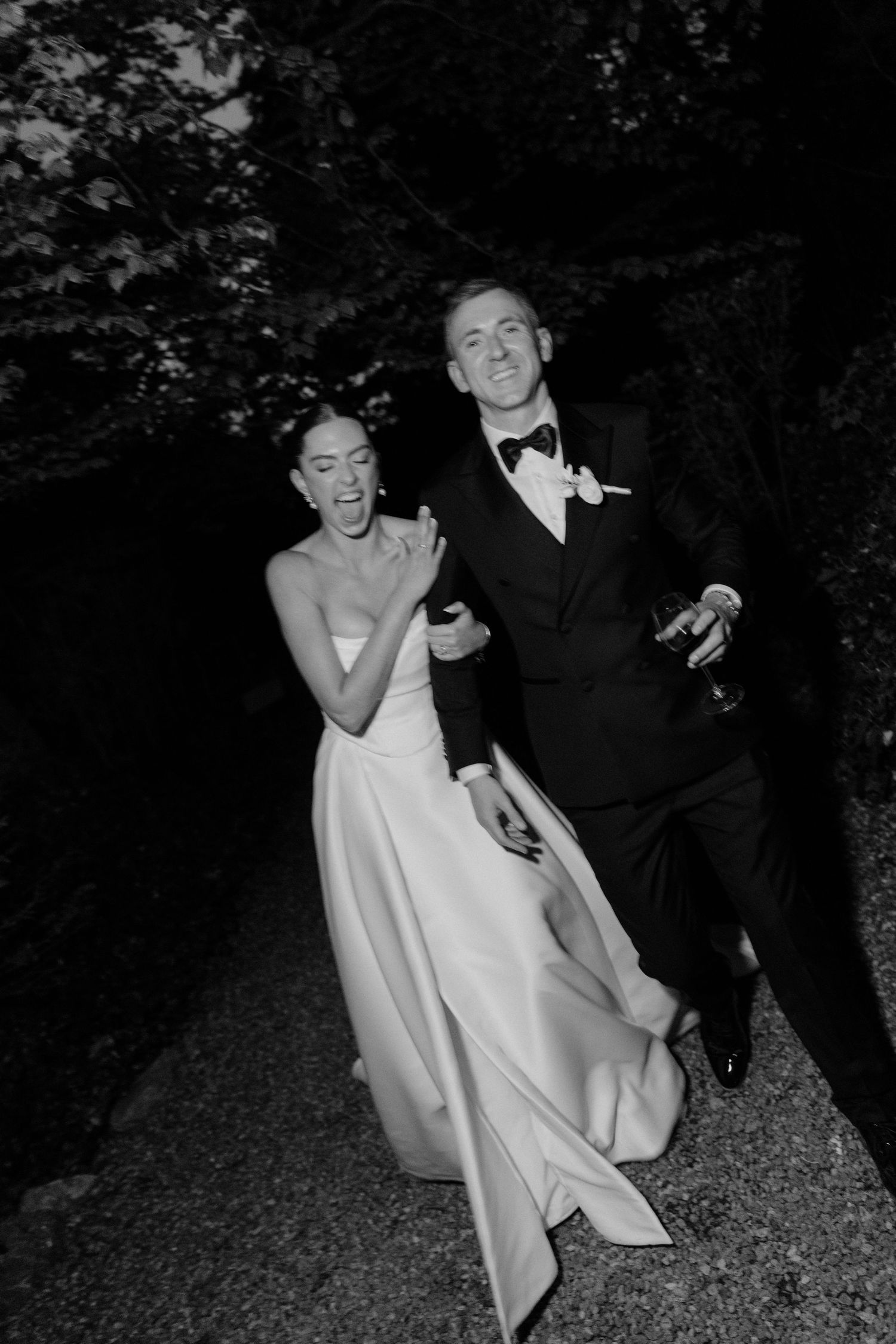 Black and white photo of a joyful bride and groom in formal attire walking together outdoors at night.