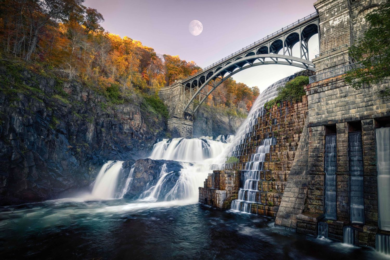 Scenic New Croton Dam waterfall with arched bridge, autumn foliage, and moon reflected in misty waters.