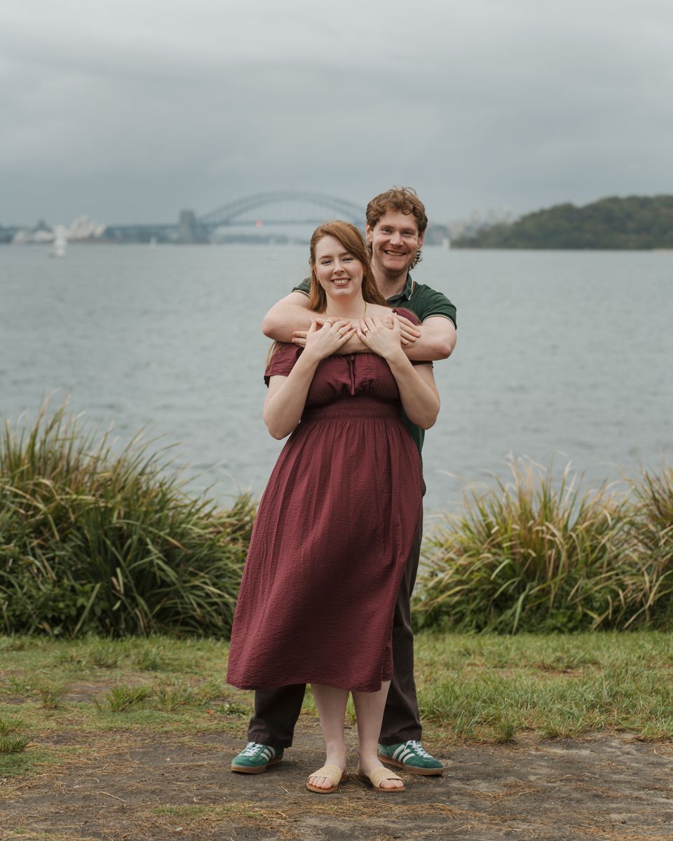 Couple hugging outdoors near Sydney Harbour Bridge, woman in burgundy dress, man in green shirt, overcast sky during engagement photoshoot
