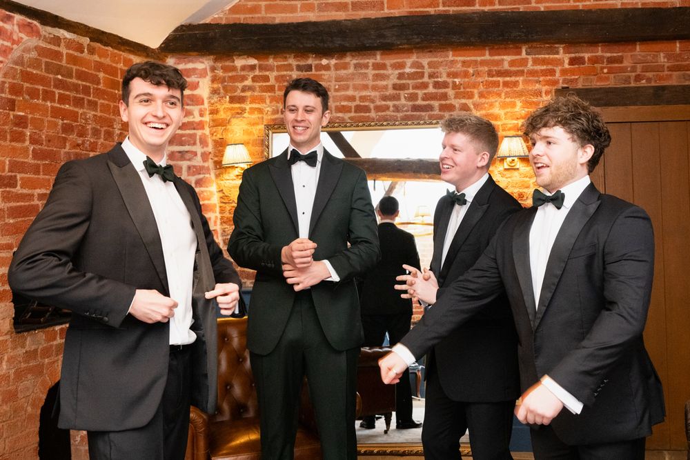Four young men in black tuxedos smiling and socializing at a formal event in a brick-walled venue.