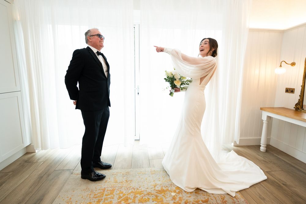 Bride in white gown pointing at groom in tuxedo during elegant first look moment indoors.