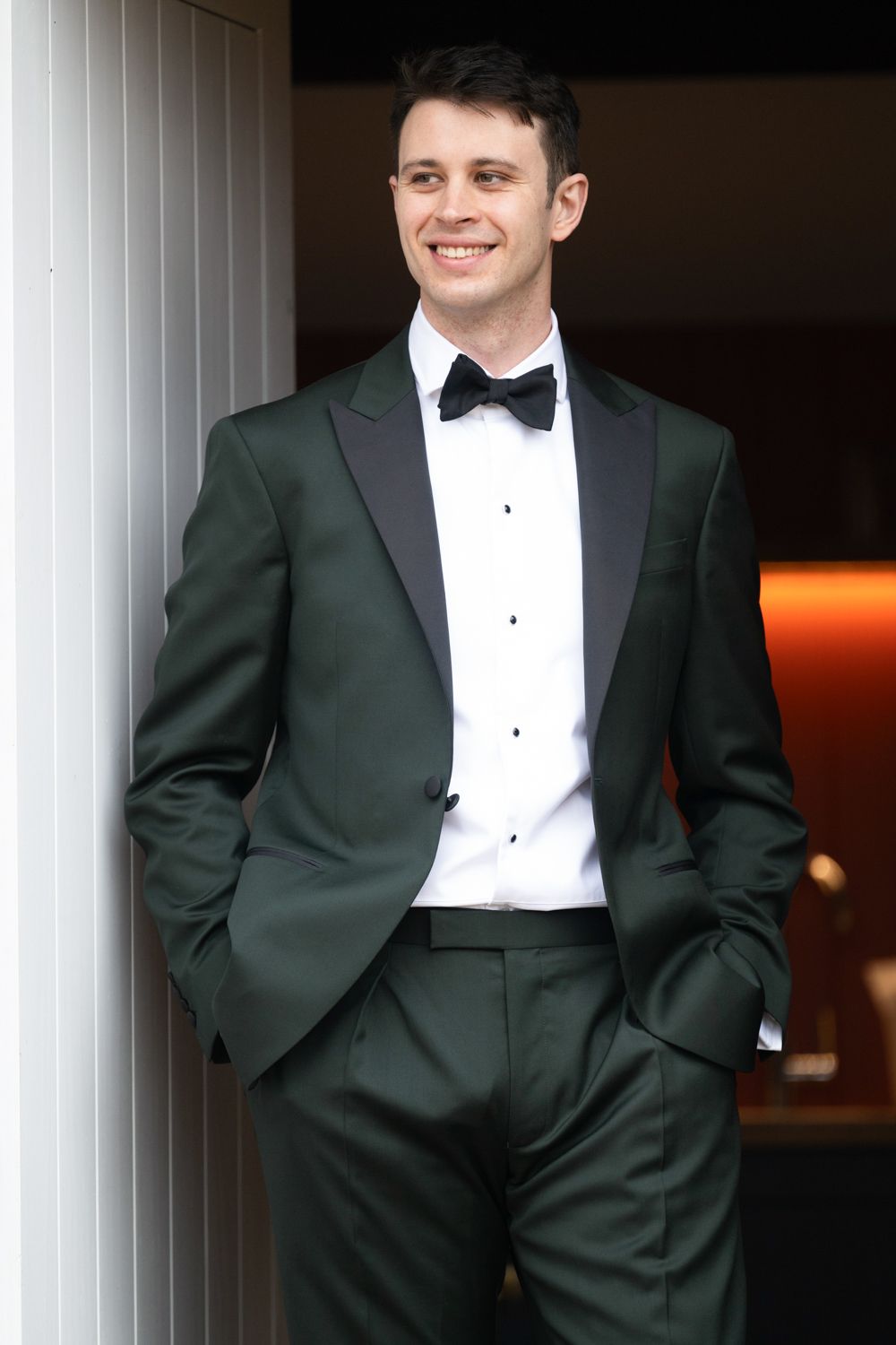 Smiling man in dark green tuxedo with black bow tie and lapels, hands in pockets, leaning against white pillar.