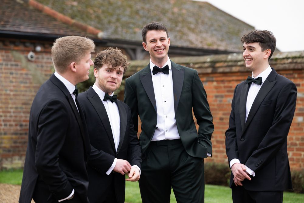 Four young men in black tuxedos and bow ties posing outdoors at a formal event.