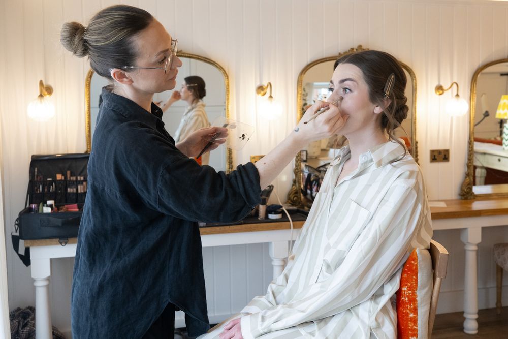 A makeup artist applying cosmetics to a bride in a bright dressing room with mirrors.