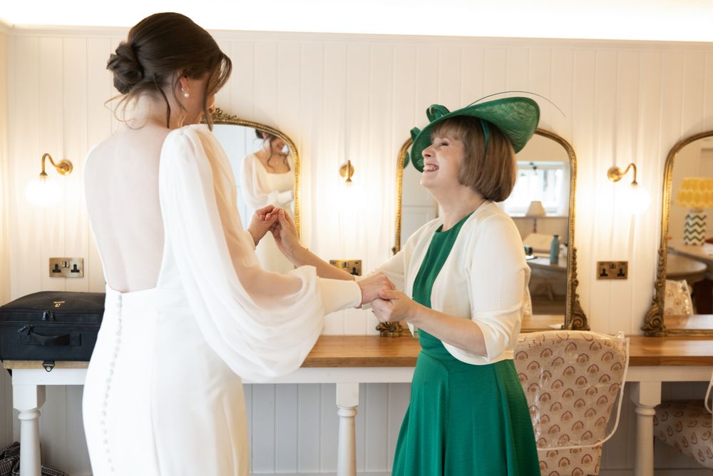 Bride in white gown holding hands with woman in green dress and hat in elegant wedding preparation room.
