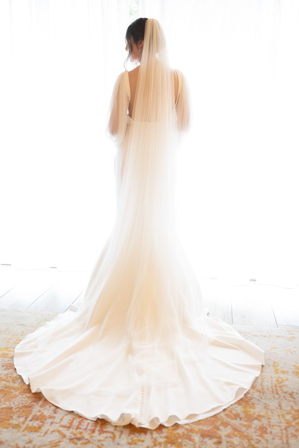 Bride in elegant white wedding gown with long veil and train standing before bright window.
