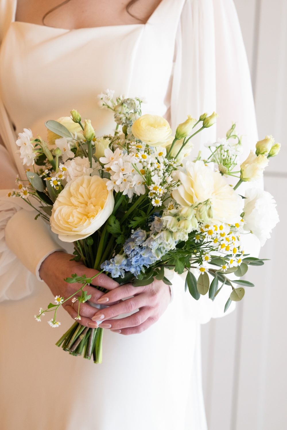 Bride holding a soft bridal bouquet with yellow roses, white lisianthus, blue hyacinths, and chamomile flowers.