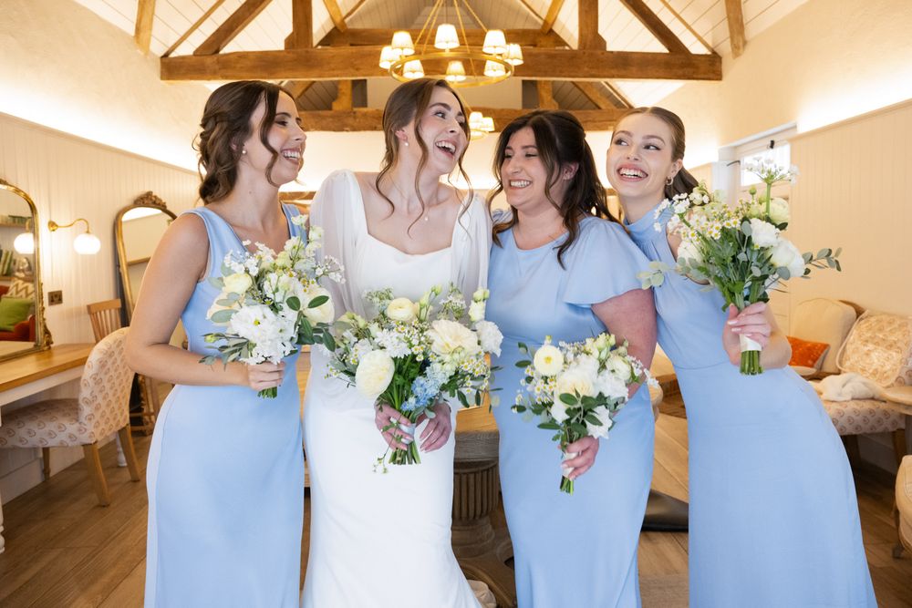 Bride and three bridesmaids in light blue dresses holding white bouquets laughing together indoors.