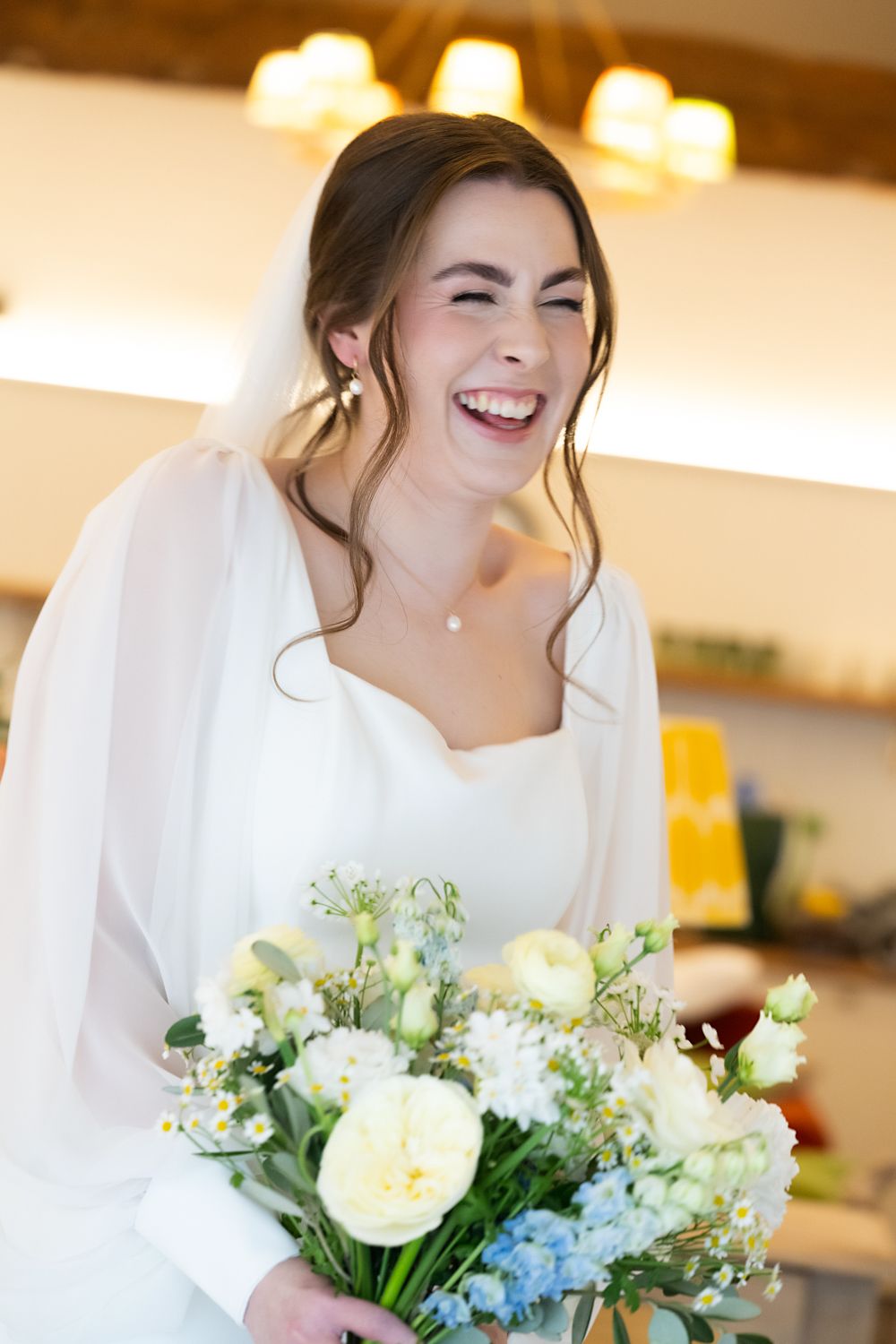 Laughing bride in white dress and veil holds bouquet of white and blue flowers indoors.