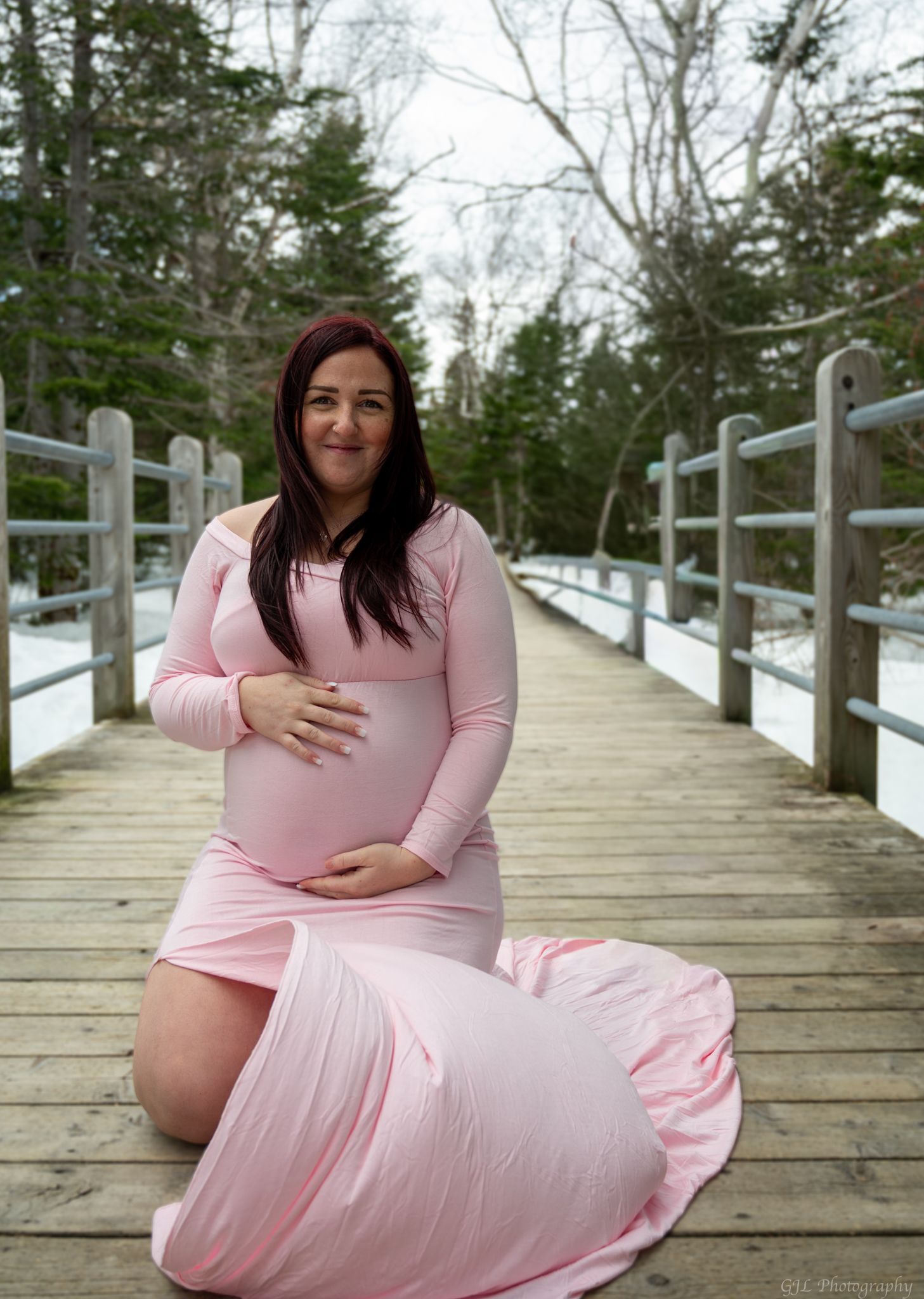 A smiling pregnant woman in a pink maternity gown poses on a wooden bridge surrounded by winter trees.