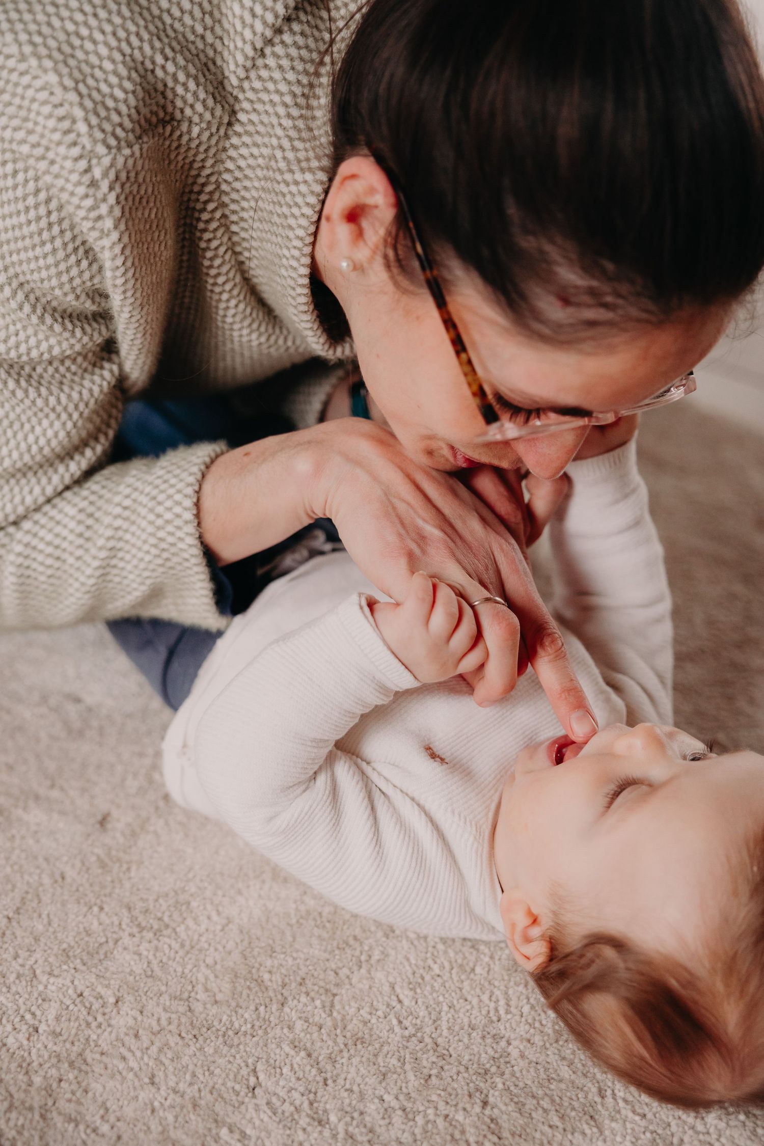 Mother with glasses leaning down to playfully interact with her smiling baby on a carpet.