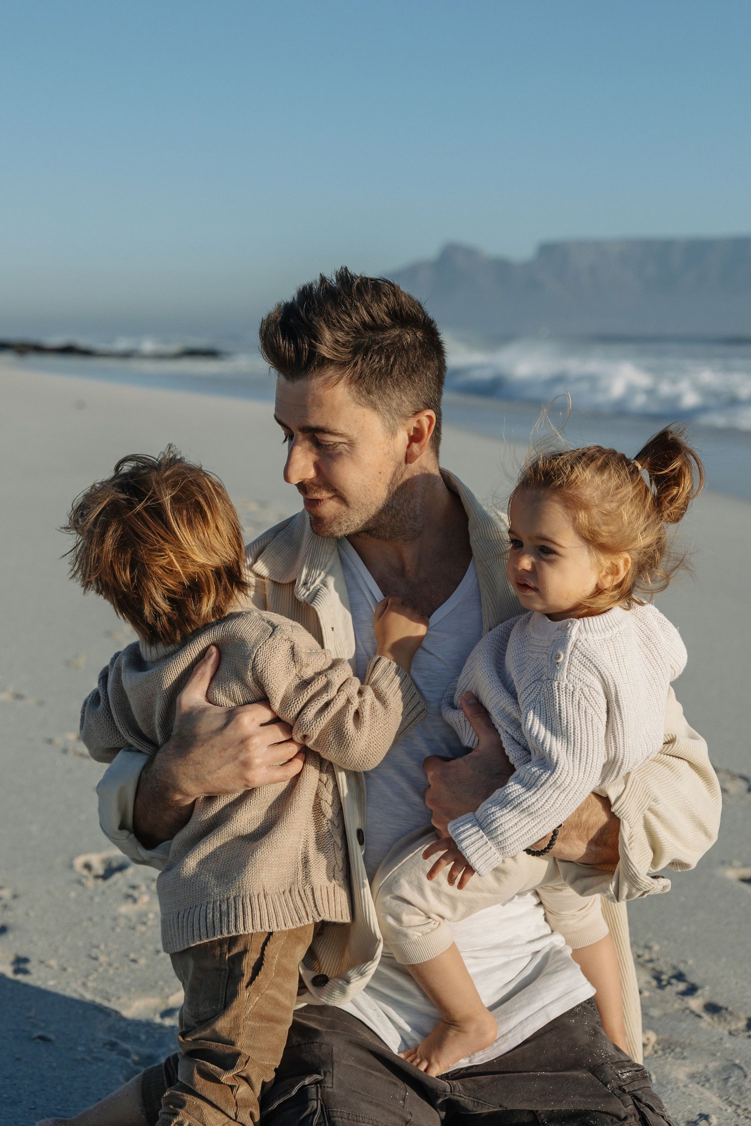 Father holds two young children on a sunny beach with ocean waves and table mountain in the background.