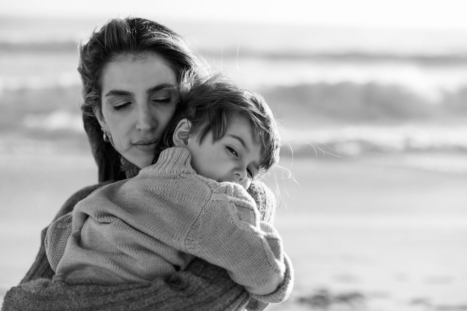 Black and white photo of a mother tenderly embracing her young child on a beach with ocean waves in the background.